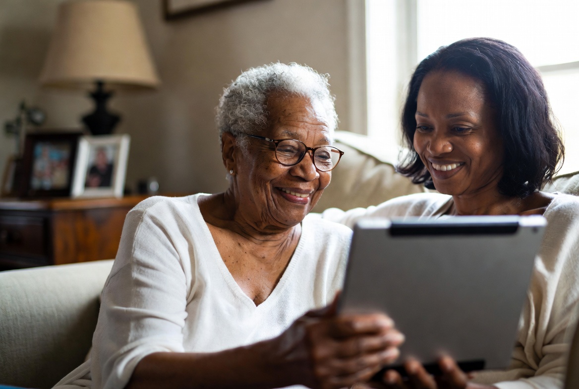 Older adult smiling during a family video call with patient tech help nearby